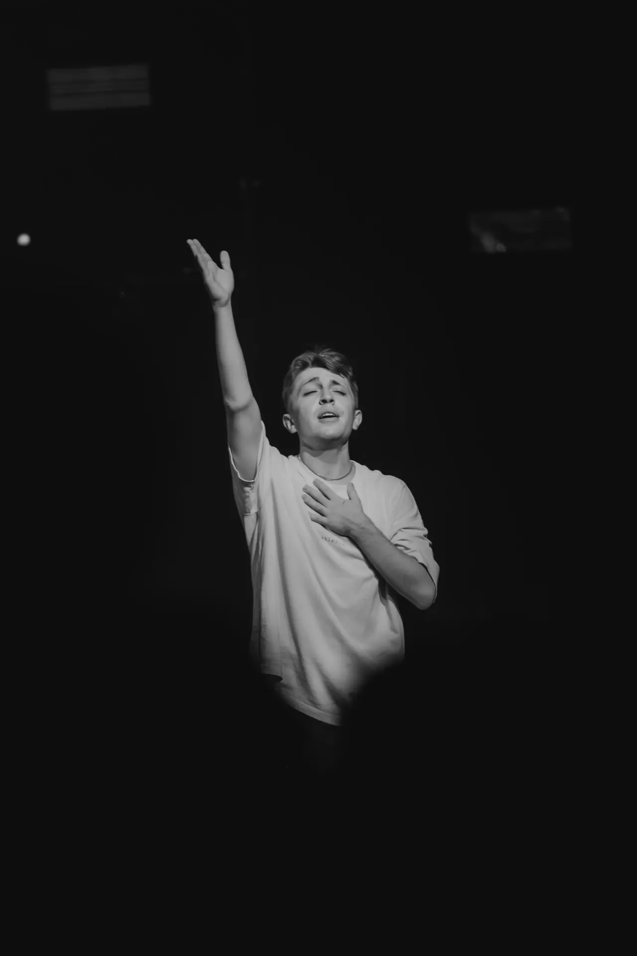 Young man worshipping with one hand raised and the other on his chest, eyes closed, in a dimly lit room.